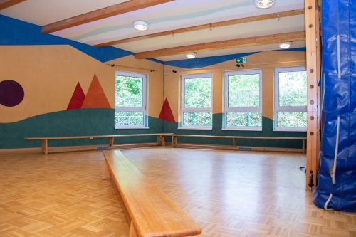 Das Bild zeigt den Bewegungsraum der Kindertagesstätte mit bunten Wandmotiven, Holzbänken und Kletterelementen.
