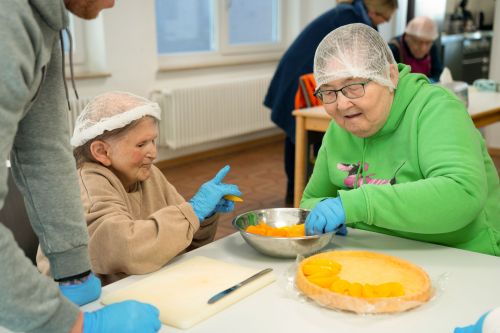 Das Bild zeigt zwei Bewohnerinnen, die gemeinsam mit Unterstützung eines Mitarbeiters in der Küche Obst für einen Kuchen vorbereiten.