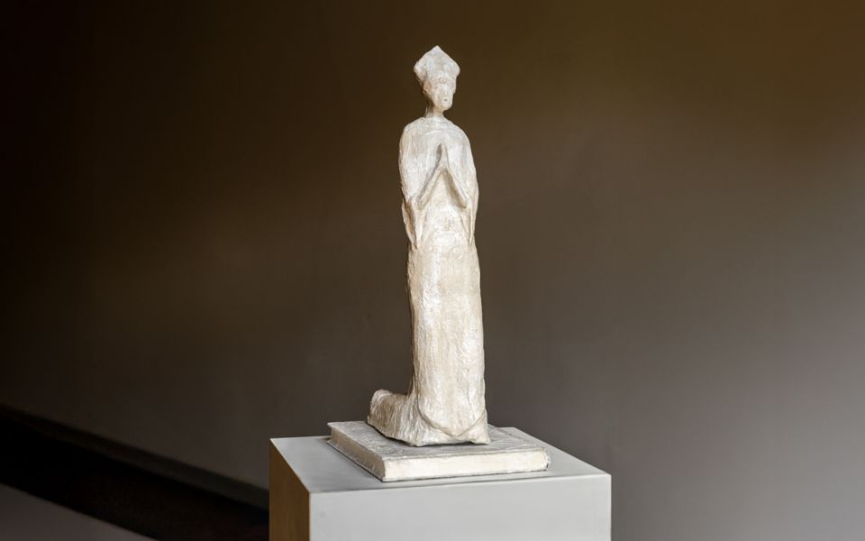 Auf dem Bild ist eine schlichte, hellfarbene Skulptur einer stehenden Figur in Gebetshaltung zu sehen, die auf einem Sockel präsentiert wird.