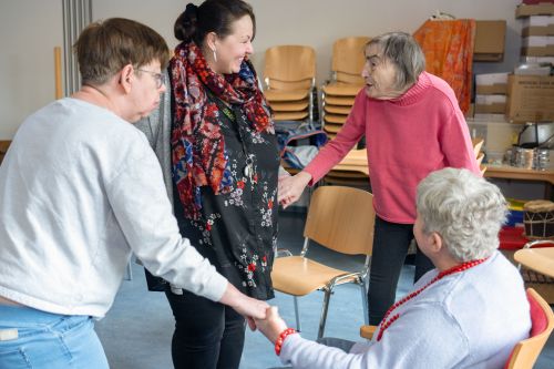 Das Bild zeigt eine Gruppe von Bewohnerinnen in einem Gemeinschaftsraum, die sich lachend an den Händen halten.