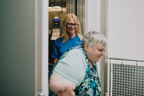 Das Bild zeigt eine Mitarbeiterin vom Pflegedienst, die einer Patientin aus dem Fahrstuhl hilft.