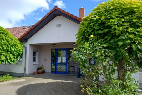 ChatGPT: Das Bild zeigt den Eingangsbereich des Wohnhauses Samuelstraße mit blauem Türrahmen, umgeben von gepflegtem Grün und einem schattenspendenden Baum.