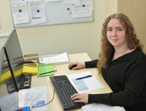 Das Bild zeigt Angelina Steiz, eine Auszubildende der Lebenshilfe Worms, wie sie lächelnd an ihrem Arbeitsplatz im Büro sitzt und am Computer arbeitet.