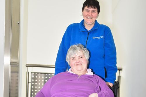 Das Bild zeigt eine Pflegerin der Lebenshilfe Worms, die gemeinsam mit einer lächelnden Bewohnerin in einem Treppenhaus unterwegs ist.
