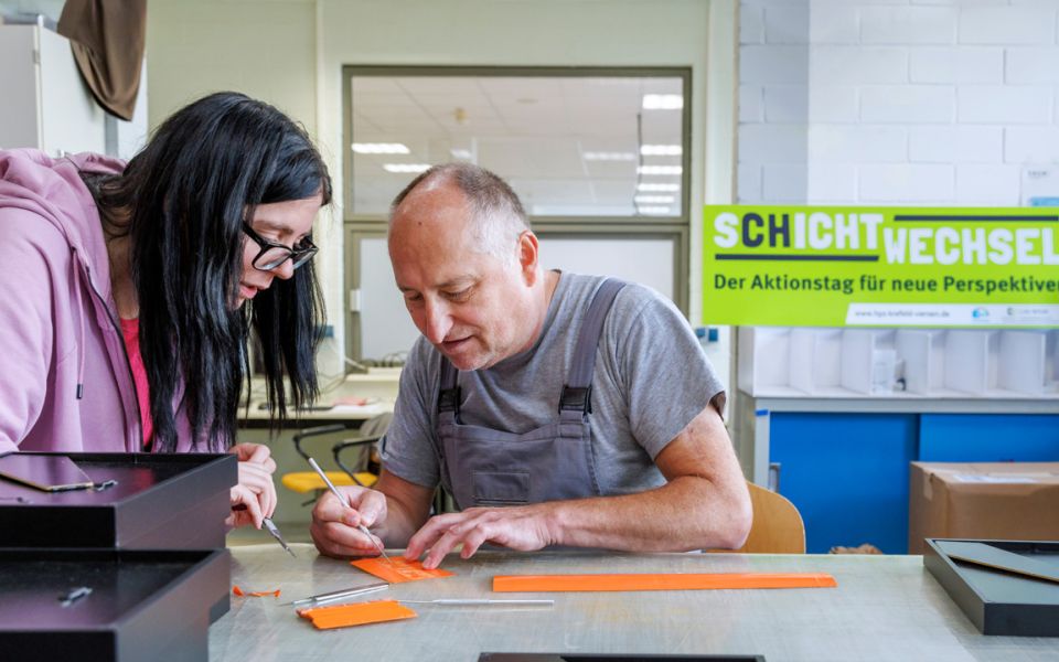 Auf dem Bild sieht man eine Frau und einen Mann, die gemeinsam in einer Werkstatt arbeiten und etwas mit einem Pinsel und einem Werkzeug basteln.