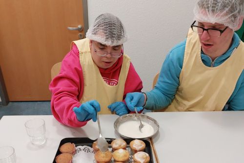 Das Bild zeigt zwei Bewohner beim gemeinsamen Verzieren von Muffins in einer Wohnküche, ausgestattet mit Schürzen, Haarnetzen und viel Freude am Tun.